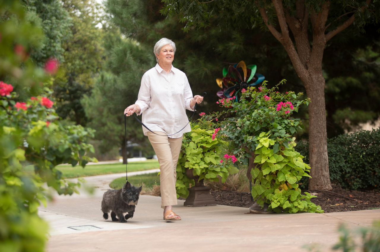 Woman walking her small dog on a path through greenery.