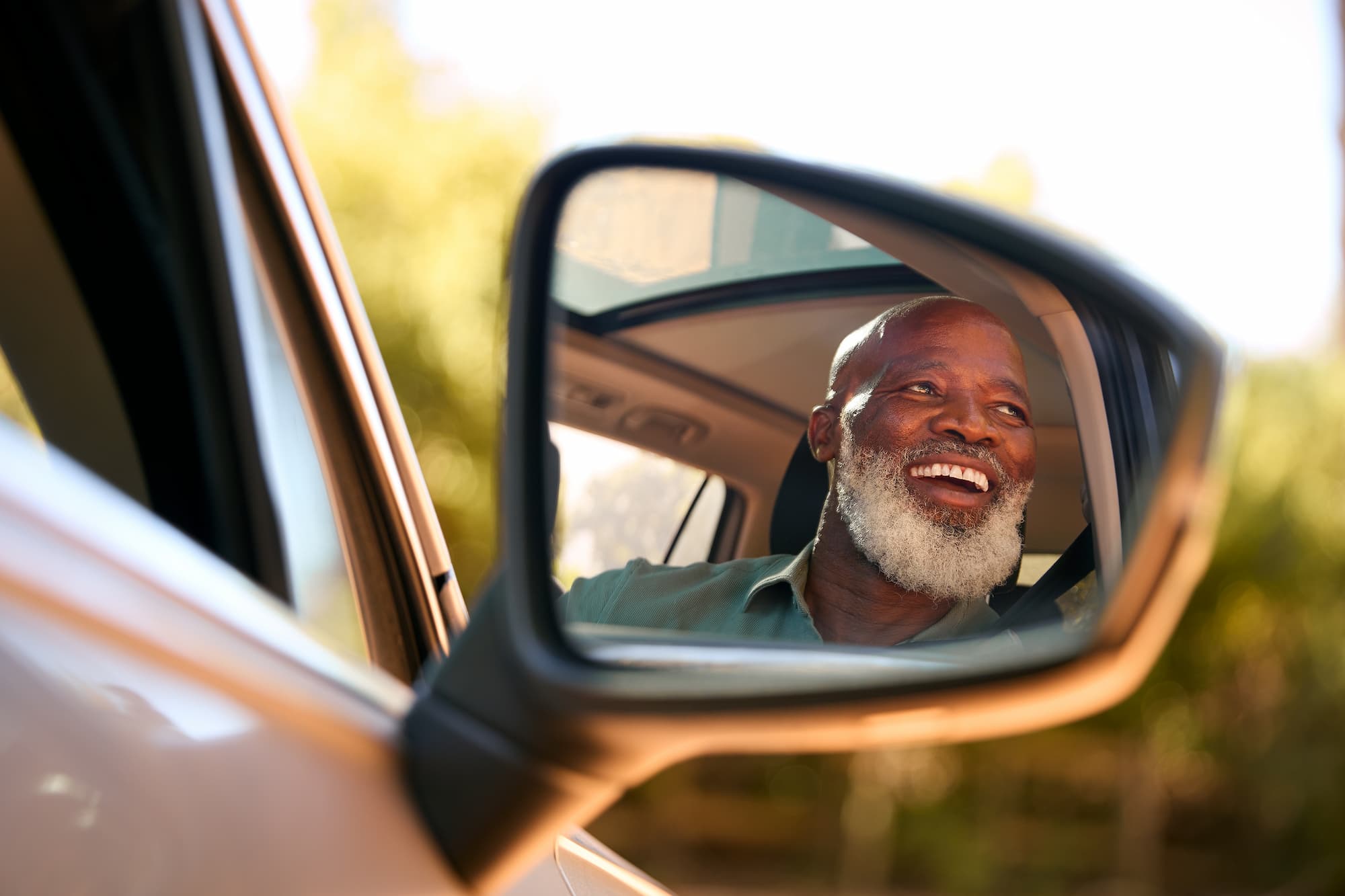 A senior male smiling in a suv car, his face appearing on the side view mirror of the car